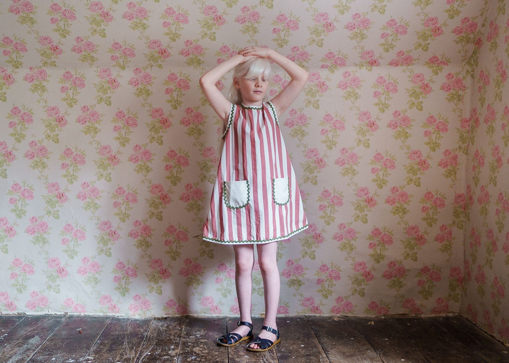 Young girl wearing a pink and white striped organic cotton dress with vintage-inspired details, standing in a floral wallpapered room, showcasing sustainable children’s clothing.