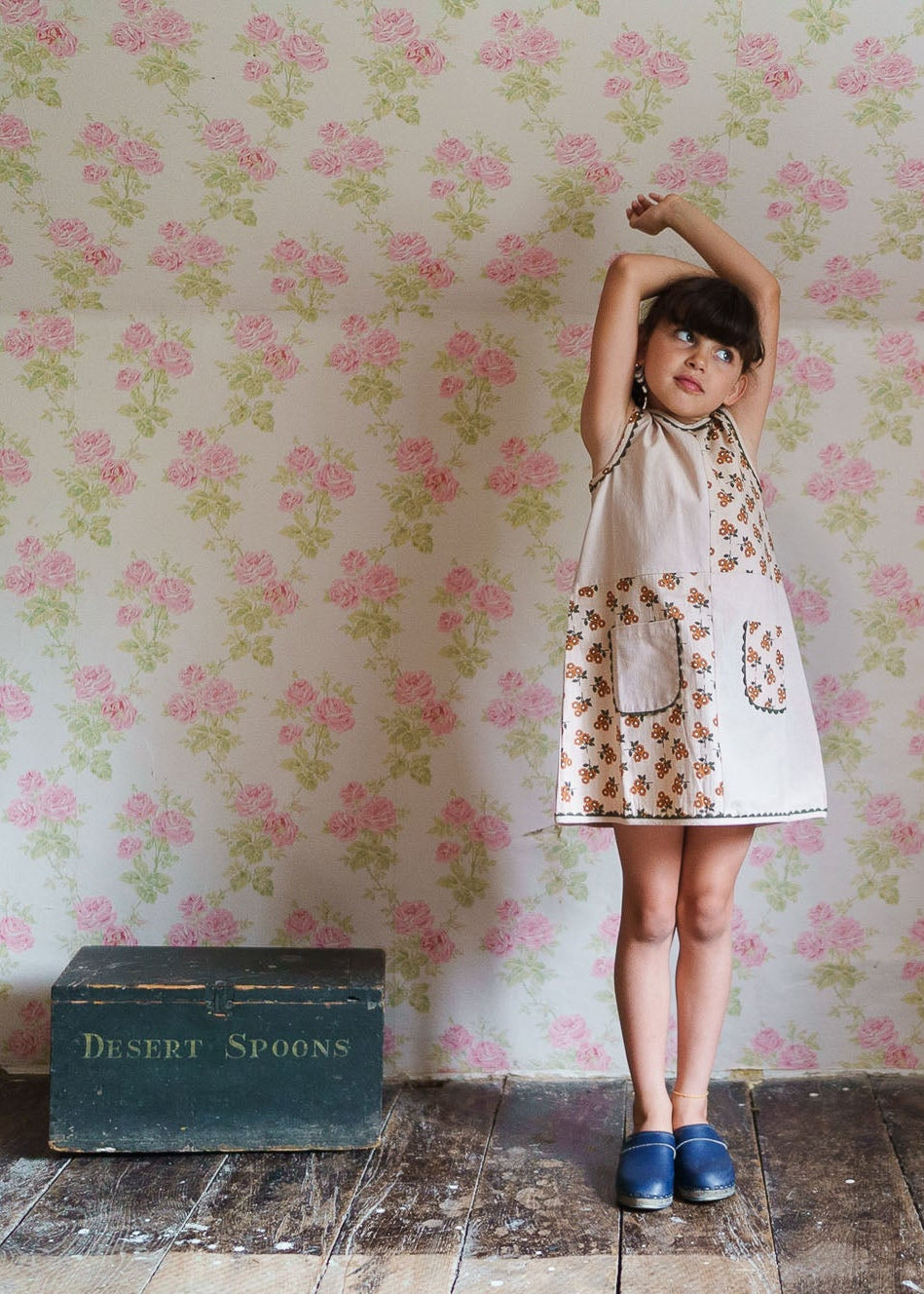 Young girl wearing a vintage-inspired organic cotton dress with floral patchwork, standing against a floral wallpaper in a sustainable children's clothing setting.