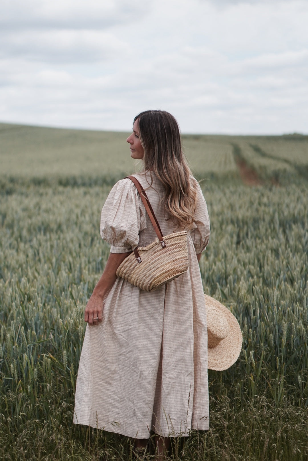 Woman in a beige dress standing in a field with a straw bag and hat.