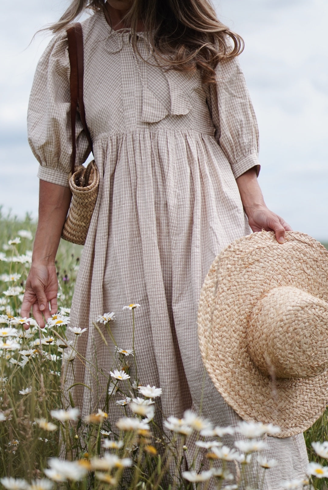 Woman in a beige checkered dress standing in a field of flowers, holding a straw hat.
