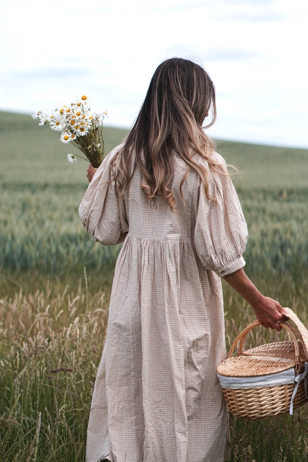 Woman in a beige dress holding flowers and a basket in a field