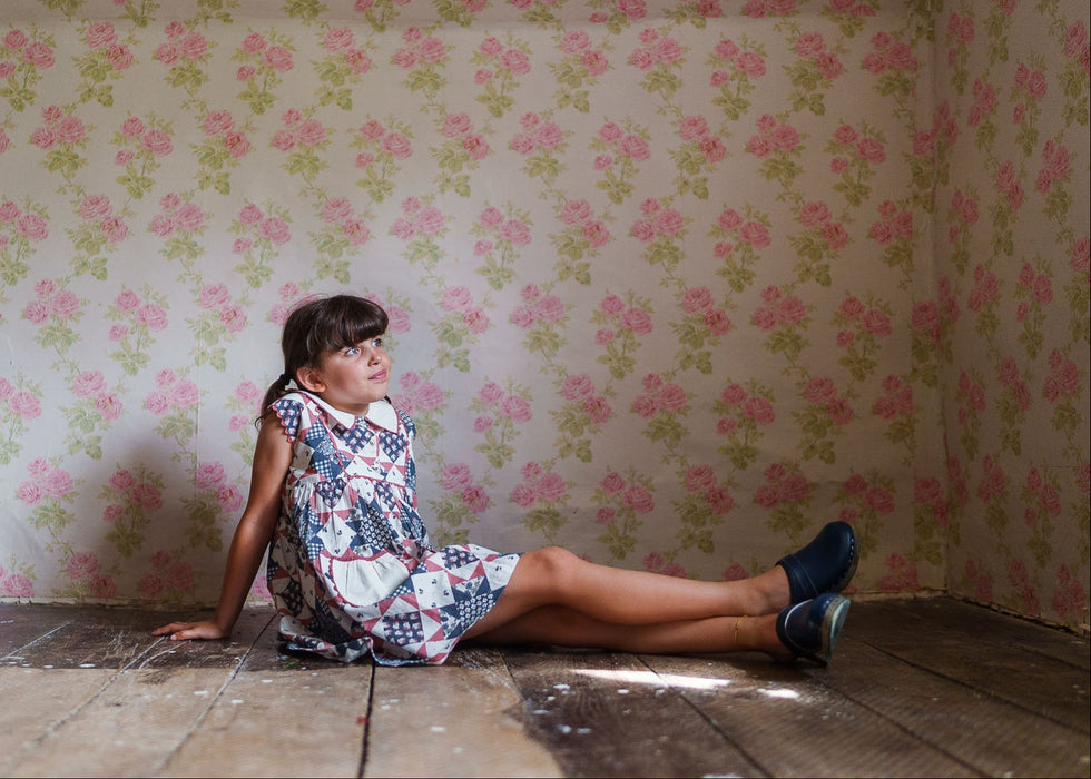 Young girl sitting on a wooden floor wearing a vintage-style organic cotton dress with blue geometric and floral patterns in a softly lit room with floral wallpaper.