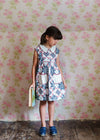 Young girl wearing a vintage-style patchwork floral dress in organic cotton, standing against a floral wallpaper background, holding a book in hand.