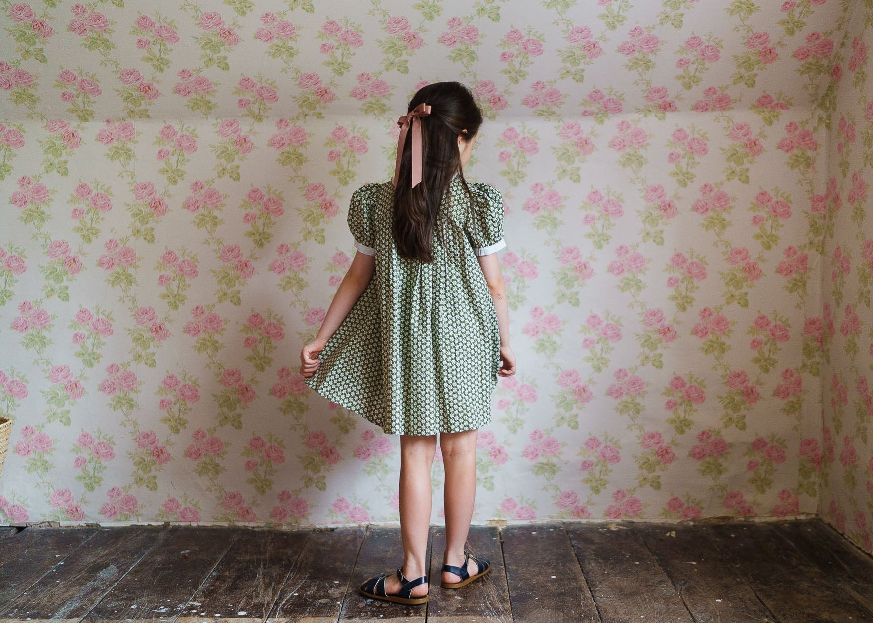 Young girl wearing a green floral vintage-style cotton blend dress with puff sleeves, standing in a room with pink floral wallpaper, showcasing sustainable children's clothing.