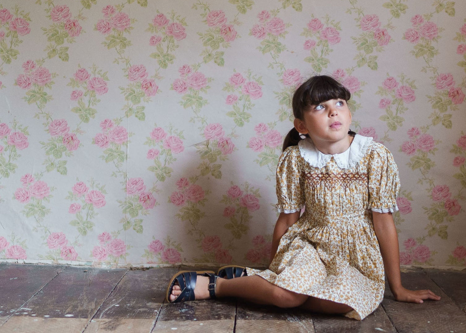 A young girl in a vintage style floral cotton blend dress with a white Peter Pan collar, sitting on a rustic wooden floor against a floral wallpaper, showcasing sustainable children’s clothing.