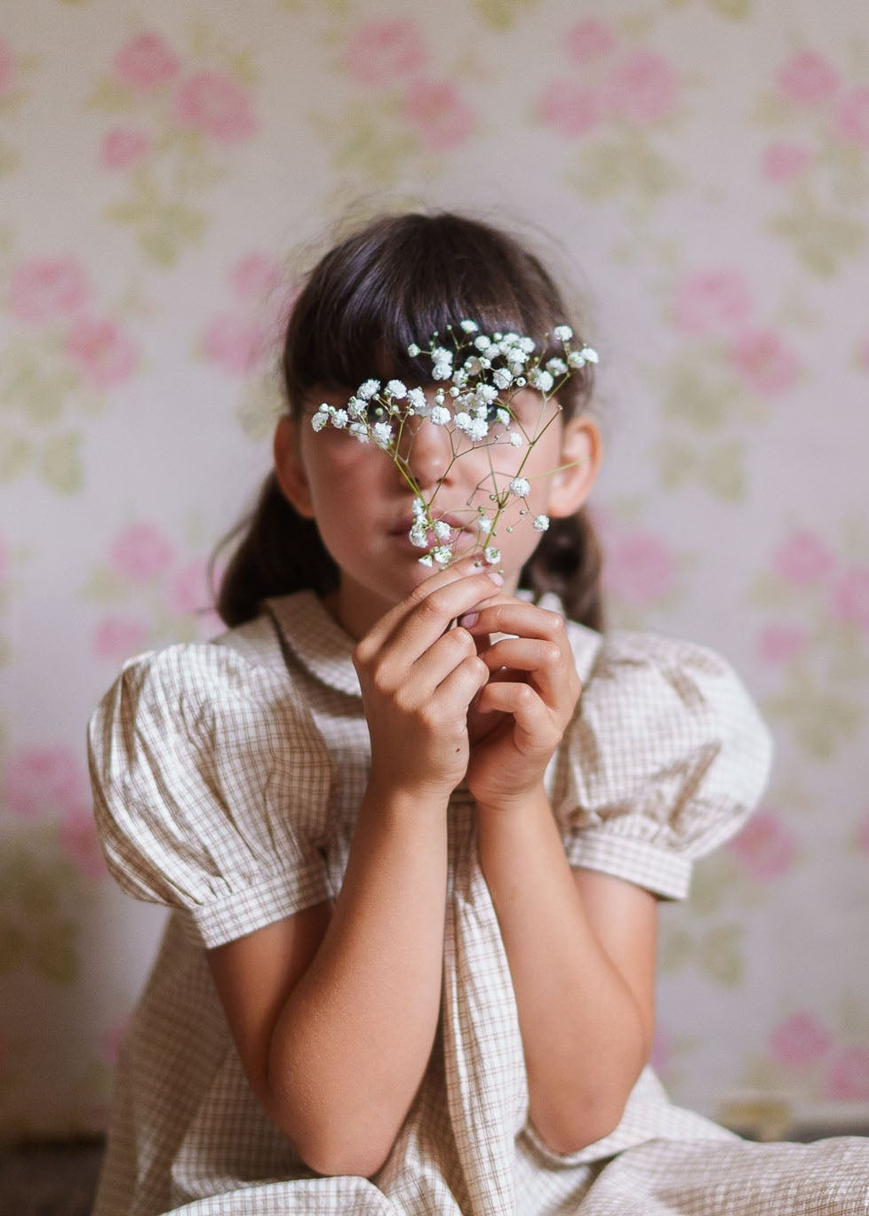 A young girl in a vintage style yellow checkered cotton dress holding delicate white flowers in front of her face, sitting against a soft floral patterned background.