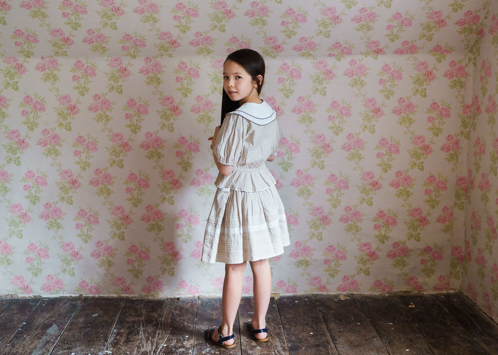 Young girl wearing a cream checkered organic cotton skirt with vintage style, standing against a floral wallpaper backdrop in a natural, ethically made children's clothing setting.