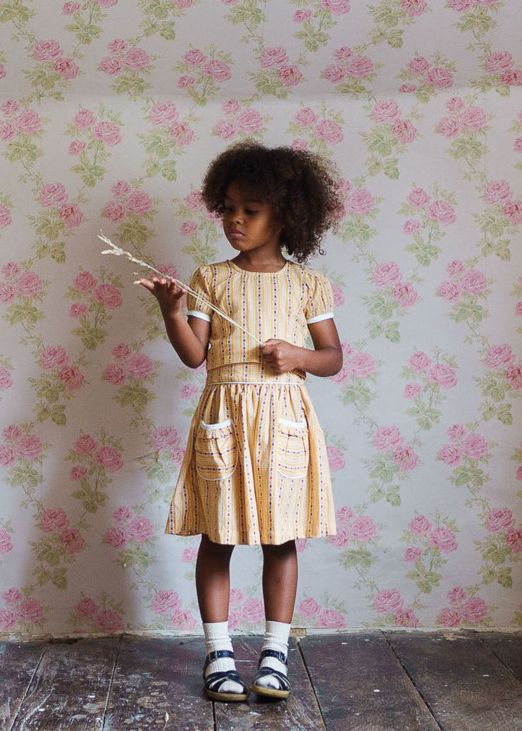 Young girl wearing a vintage style yellow cotton dress with pockets, standing against floral wallpaper, embodying slow, natural, and ethical children's clothing.