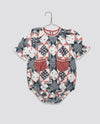 Vintage style baby romper with organic cotton patchwork floral print in muted red and blue tones, featuring puff sleeves and small front pockets in natural materials.