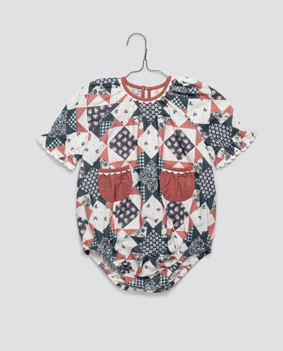 Vintage style baby romper with organic cotton patchwork floral print in muted red and blue tones, featuring puff sleeves and small front pockets in natural materials.