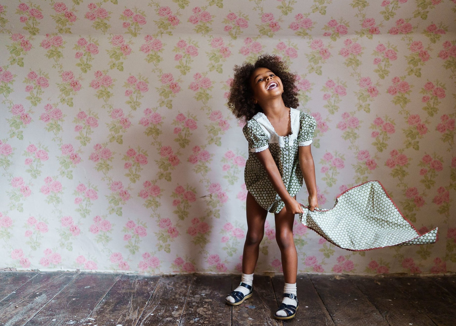 Happy young girl wearing a vintage-style green romper made from organic cotton, playing indoors on rustic wooden flooring with floral wallpaper in the background.
