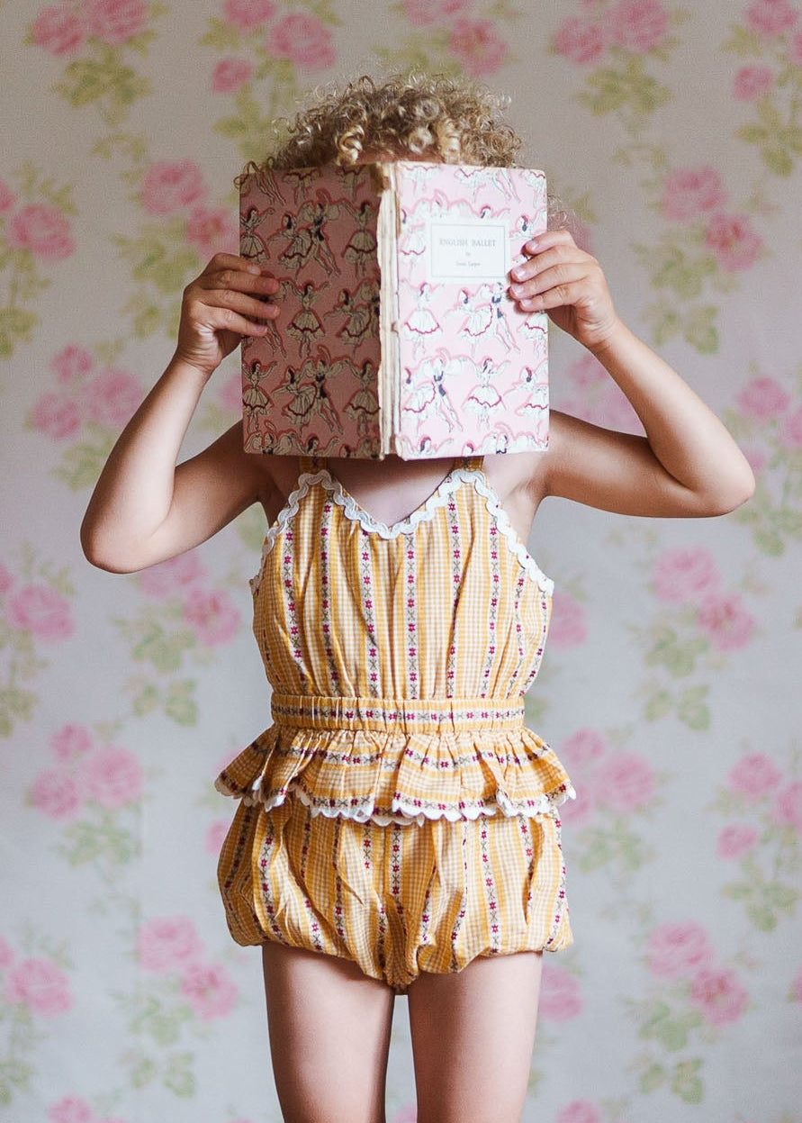 Child wearing a vintage style yellow floral romper with delicate lace trim, made from organic cotton, standing against a soft floral wallpaper background while holding a patterned book in front of their face.
