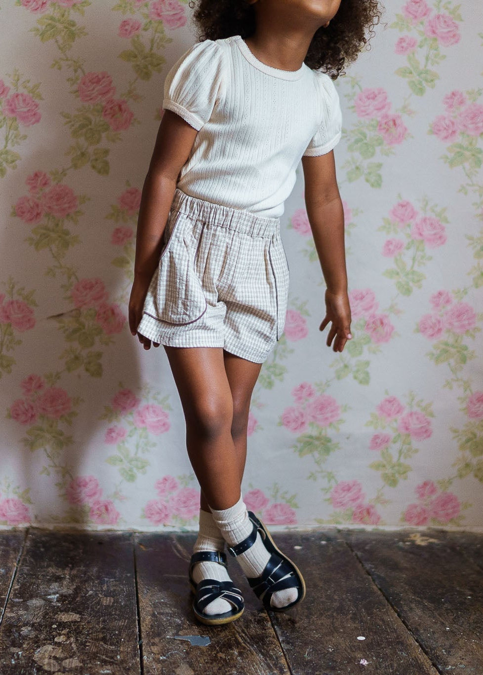 Young child wearing vintage-inspired organic cotton shorts in a soft check pattern paired with a white textured top, standing on rustic wooden flooring.