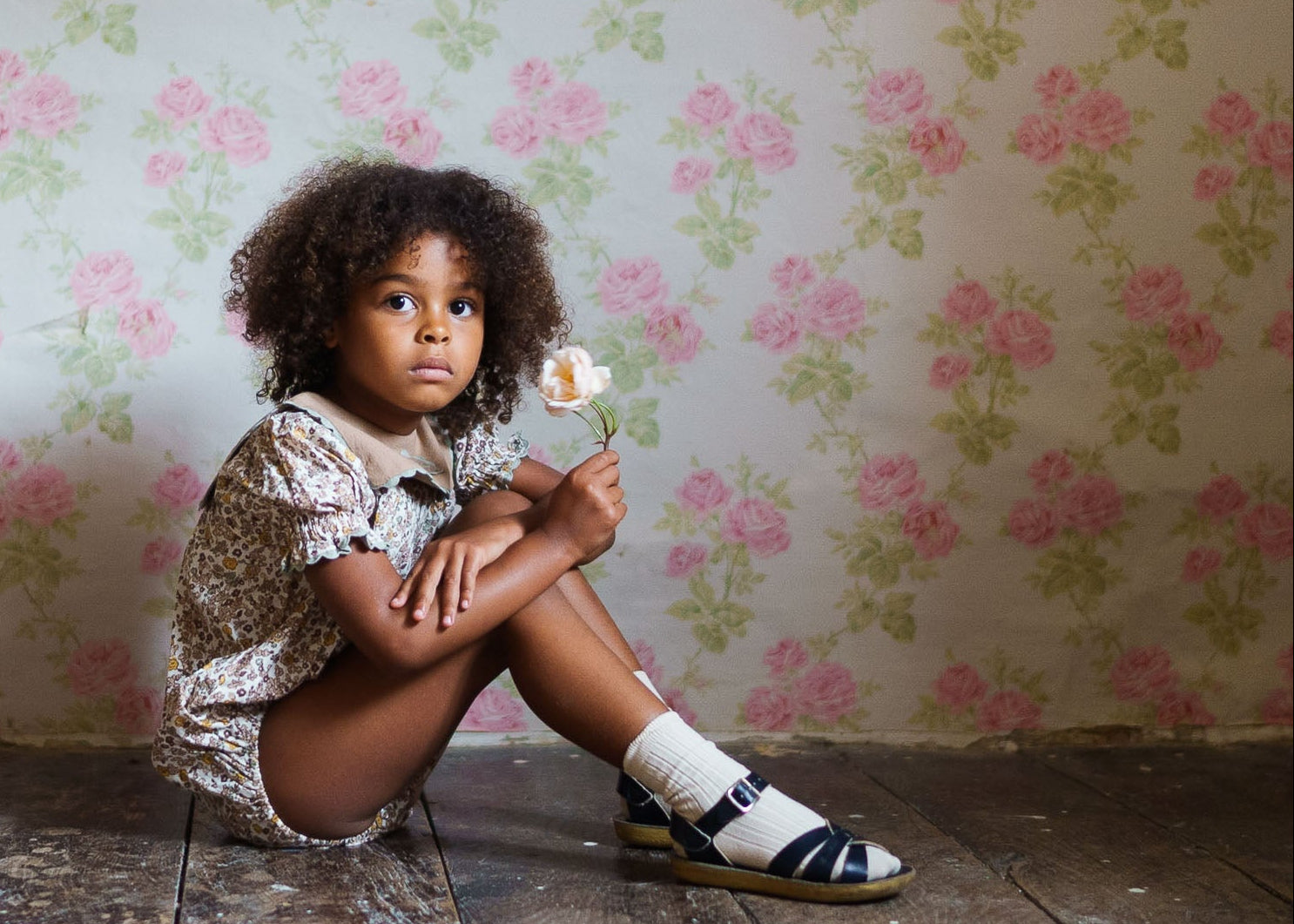 Young child wearing organic cotton floral romper sitting on wooden floor holding a flower, styled in vintage-inspired sustainable children's clothing.