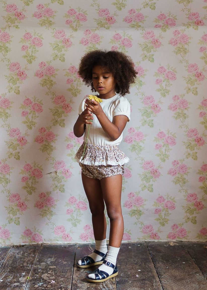 Young girl wearing a cream organic cotton t-shirt and vintage-style floral shorts, standing on a rustic wooden floor with pink floral wallpaper, holding a yellow flower.