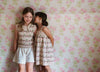Two young girls wearing vintage style children's clothing made from organic cotton, one in a cherry patterned hand-knitted vest and the other in a matching dress, standing against a floral wallpaper.
