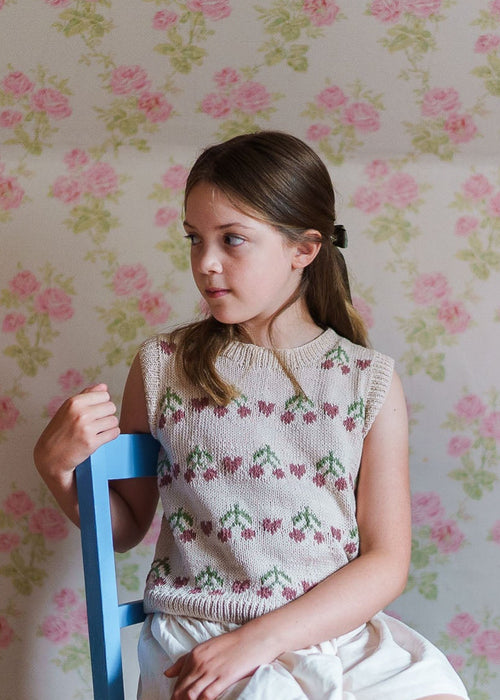 Young girl wearing a vintage style hand-knitted cotton vest with cherry patterns, sitting on a blue chair against a floral wallpaper background, showcasing sustainable children's clothing.