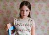 Young girl wearing a vintage-style hand-knitted cotton vest with cherry patterns, sitting on a blue chair against a floral wallpaper background, reflecting natural and sustainable children’s clothing.