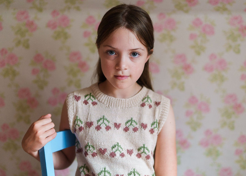 Young girl wearing a vintage-style hand-knitted cotton vest with cherry patterns, sitting on a blue chair against a floral wallpaper background, reflecting natural and sustainable children’s clothing.