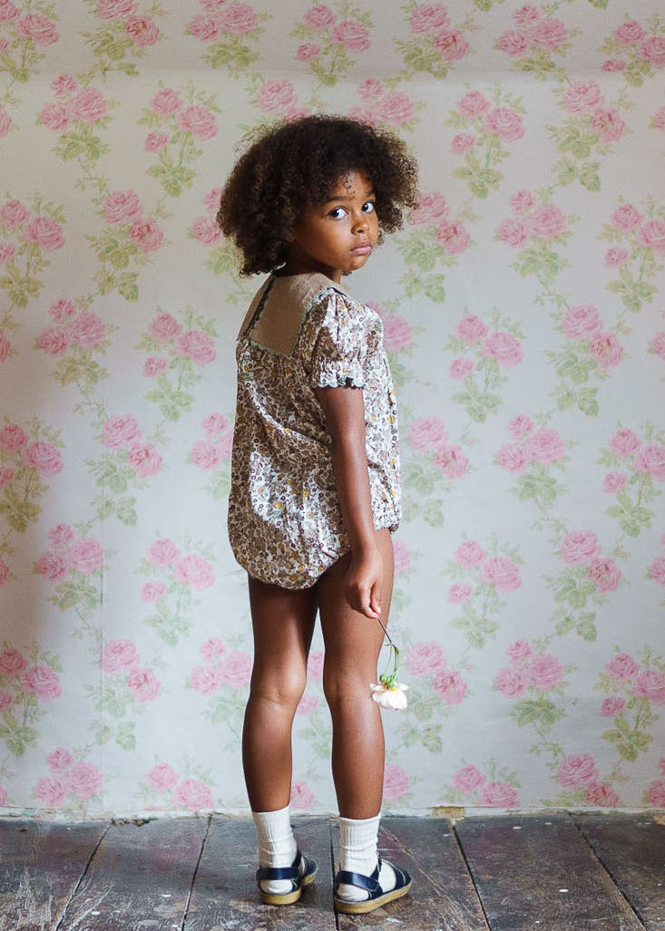 Young child wearing a vintage style organic cotton floral romper, standing barefoot on rustic wooden floor against a floral wallpaper backdrop, holding a small flower.