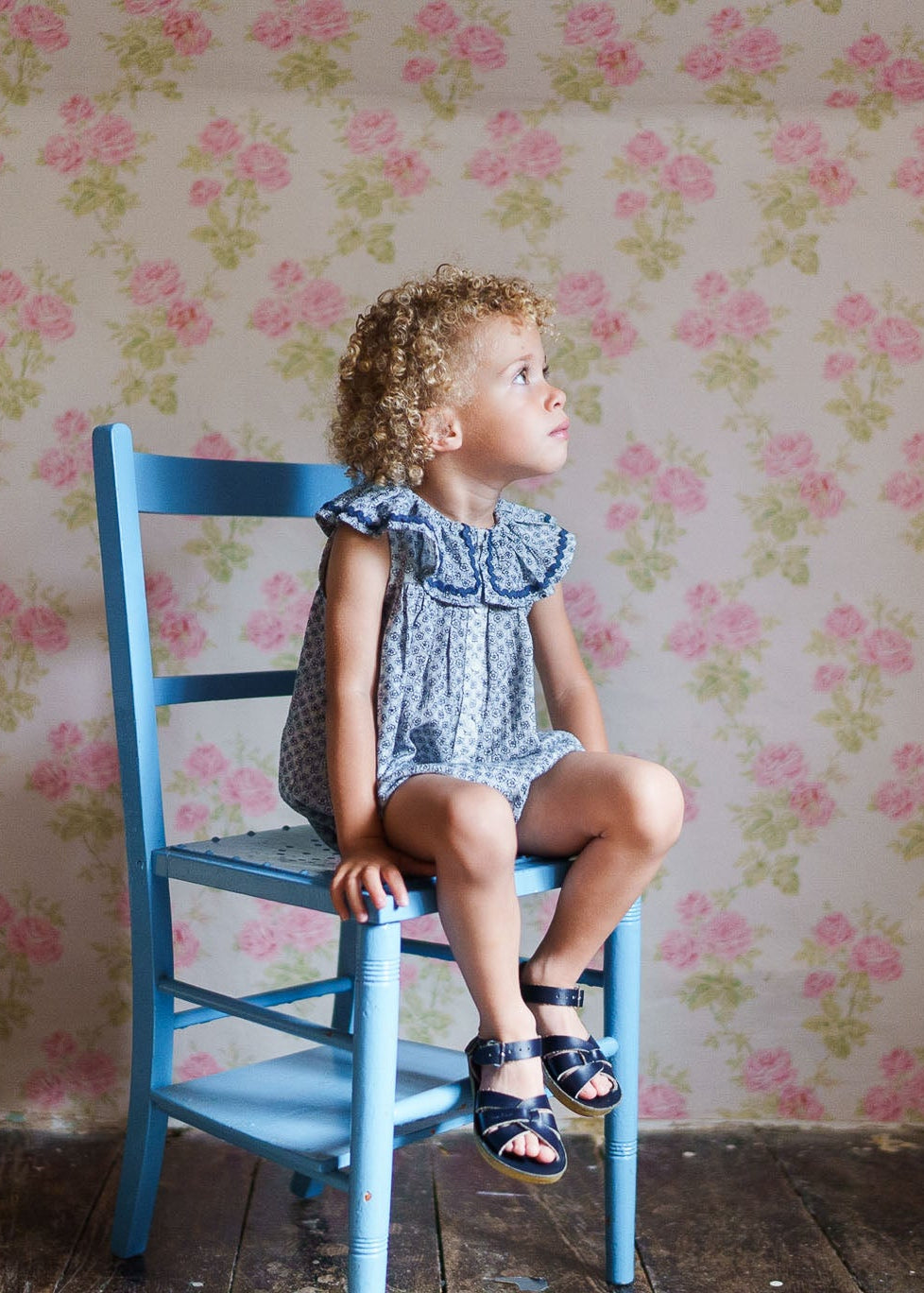 Young child sitting on a blue wooden chair wearing a vintage-style, organic cotton romper with delicate floral patterns, set against a soft floral wallpaper background.