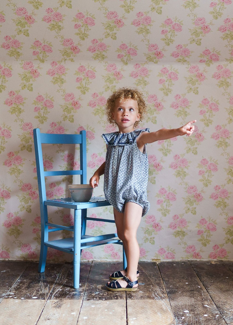 Young child wearing a vintage style romper with a subtle floral pattern, standing beside a small blue wooden chair in a room with pink rose wallpaper, showcasing natural and organic children’s clothing.
