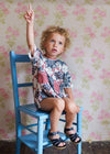 Young child sitting on a blue wooden chair wearing a vintage style romper in soft floral patchwork, showcasing organic, ethically made children's clothing in a cozy, natural setting.