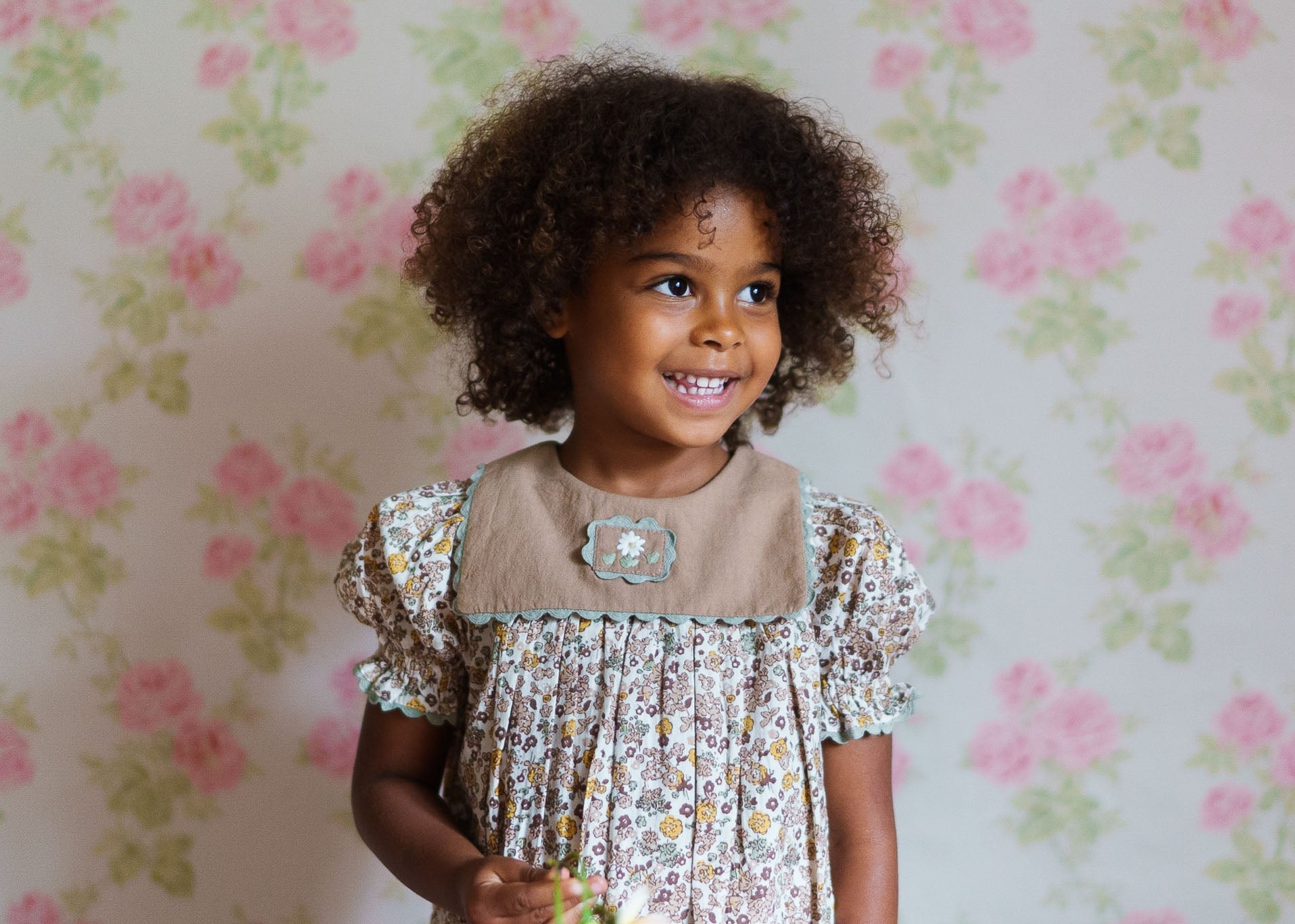 Smiling toddler girl wearing a vintage-style floral baby romper made from organic cotton, standing against a soft pink and green floral wallpaper background.