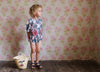 Young child wearing a vintage-style patchwork floral romper made from soft organic cotton, standing on wooden floor with a woven basket nearby, in a cozy, natural setting.