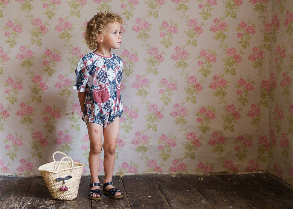 Young child wearing a vintage-style patchwork floral romper made from soft organic cotton, standing on wooden floor with a woven basket nearby, in a cozy, natural setting.