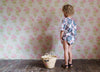 Toddler wearing a natural cotton romper with a patchwork floral pattern, standing on a rustic wooden floor next to a woven basket, in a vintage-style room.