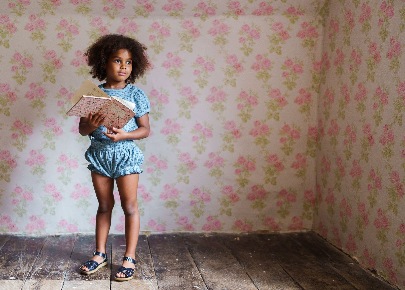 Young girl wearing a vintage style organic cotton outfit with floral print, standing on rustic wooden floor against a pink floral wallpaper, holding an open book.