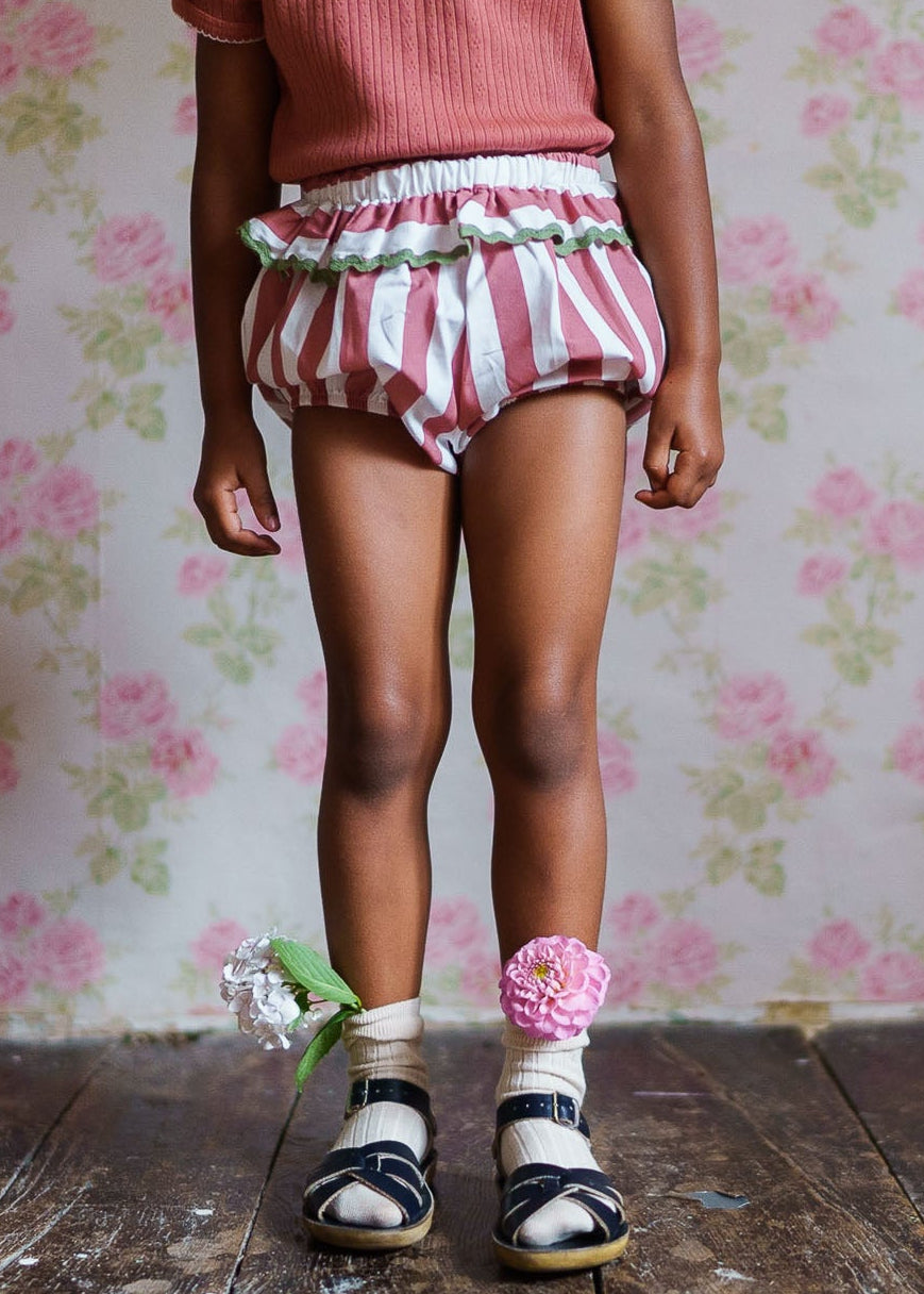 Child wearing vintage style raspberry striped bloomers made from organic cotton, paired with socks decorated with fabric flowers and black sandals, standing on wooden floor.