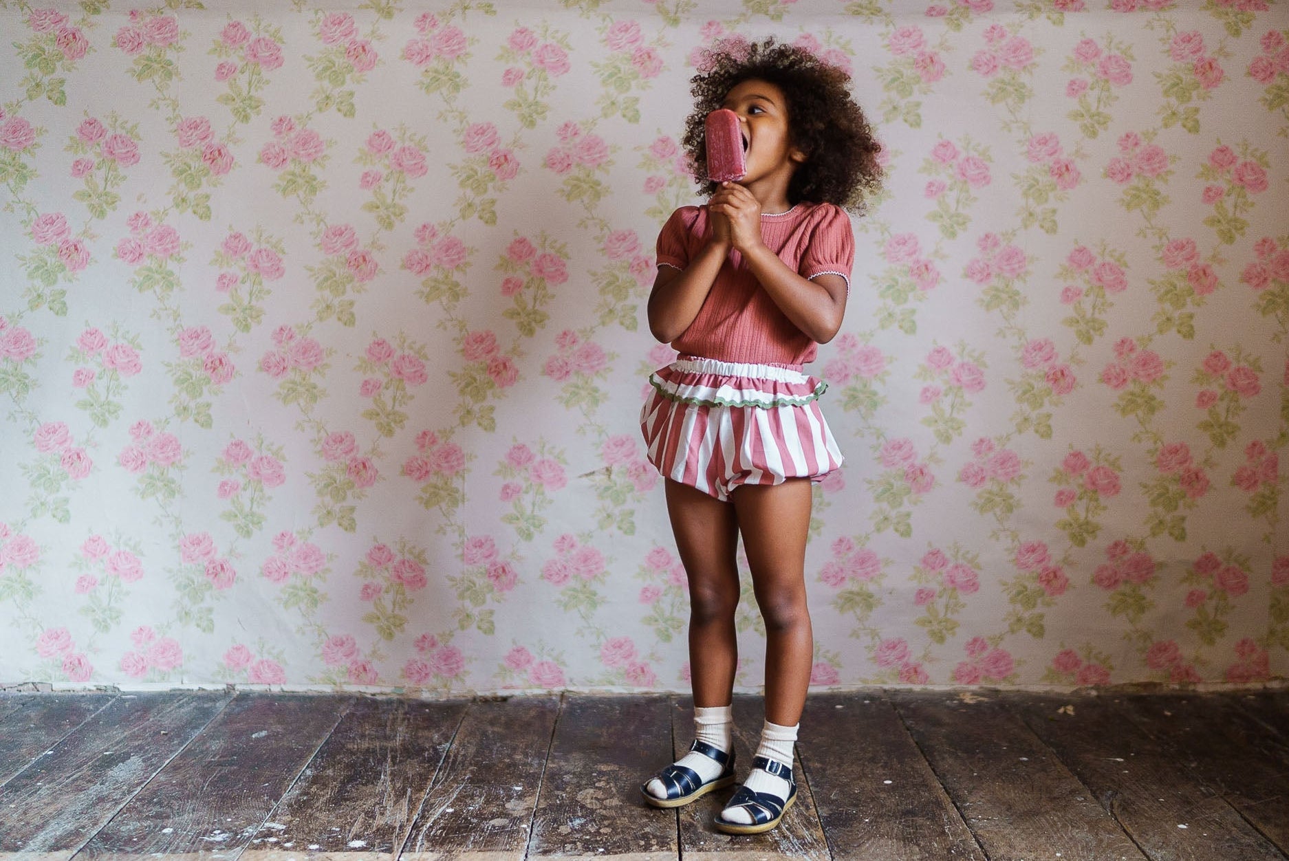 Toddler girl wearing vintage style organic cotton frilly bloomers in pink raspberry stripes enjoying a natural popsicle against a floral wallpaper background.