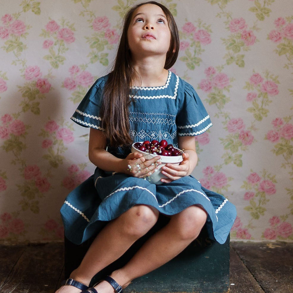 Young girl sitting on a wooden stool against a floral wallpapered wall.
