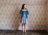 Young girl wearing a blue organic cotton jacket with vintage style dress standing against a floral wallpaper background in a natural, ethically made children's clothing setting.
