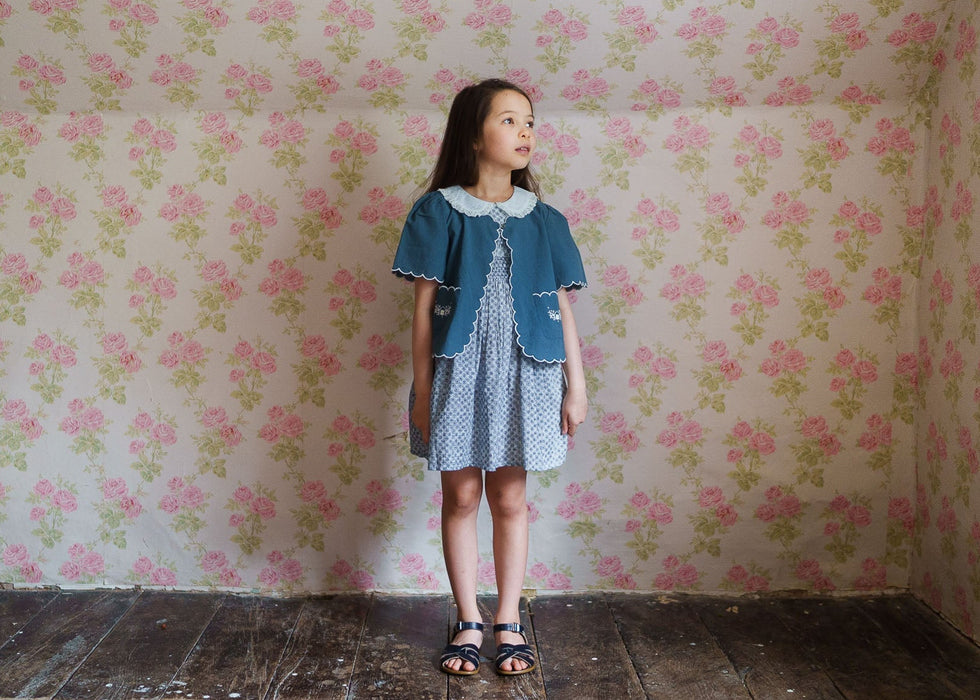 Young girl wearing a blue organic cotton jacket with vintage style dress standing against a floral wallpaper background in a natural, ethically made children's clothing setting.
