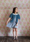 Girl wearing vintage style organic cotton blue jacket with scalloped edges over a patterned dress, standing against floral wallpaper in a natural, ethical children’s clothing setting.