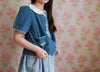 Child wearing a vintage style blue organic cotton jacket with scalloped edges and delicate white embroidery over a patterned dress, standing against floral wallpaper.