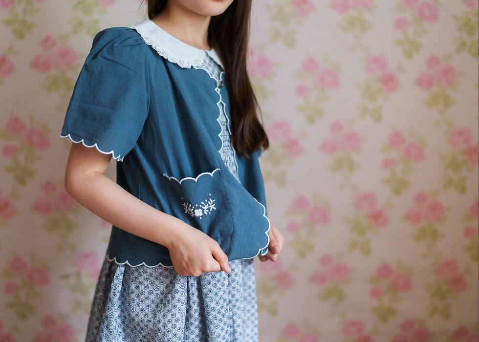 Child wearing a vintage style blue organic cotton jacket with scalloped edges and delicate white embroidery over a patterned dress, standing against floral wallpaper.