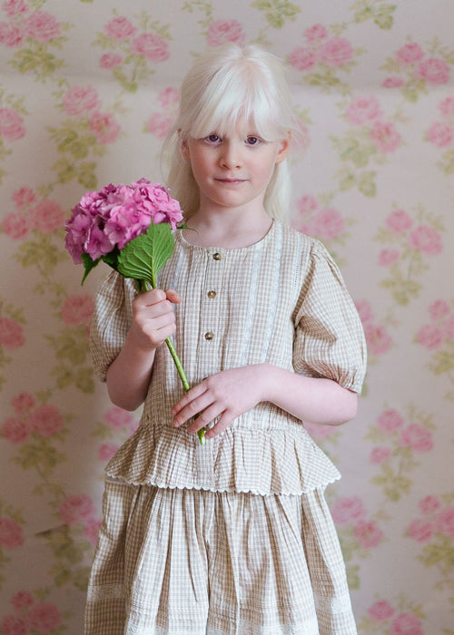 Young girl with pale hair wearing a soft straw check cotton blouse holding a pink flower, set against a delicate floral vintage-style background, showcasing sustainable children’s clothing.