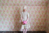 Young girl wearing a vintage-style, straw check blouse and skirt made from natural organic cotton, standing against a floral wallpaper holding pink flowers with a calm expression.