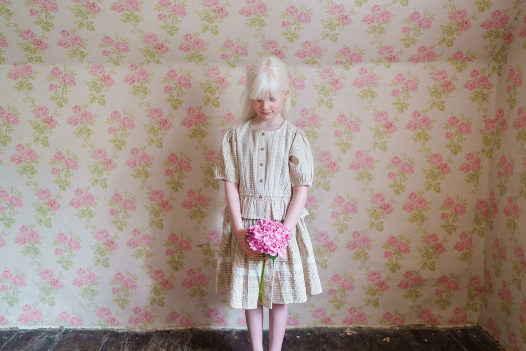 Young girl wearing a vintage-style, straw check blouse and skirt made from natural organic cotton, standing against a floral wallpaper holding pink flowers with a calm expression.