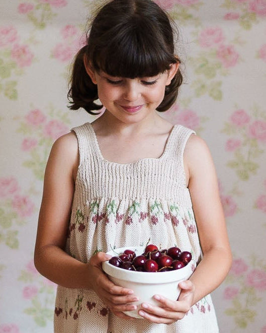 Young girl wearing a hand-knitted pinafore dress made from organic cotton, holding a bowl of fresh cherries against a vintage floral wallpaper background, embodying natural and ethical children's clothing.