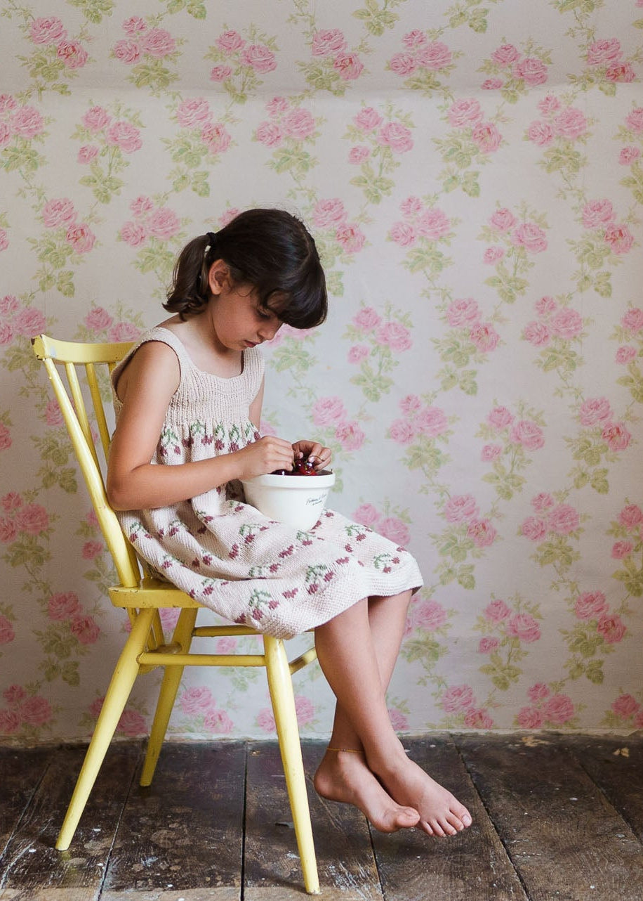 Young girl sitting barefoot on a yellow chair wearing a hand-knitted pink and cream pinafore dress with a cherry pattern, against a vintage floral wallpaper background.
