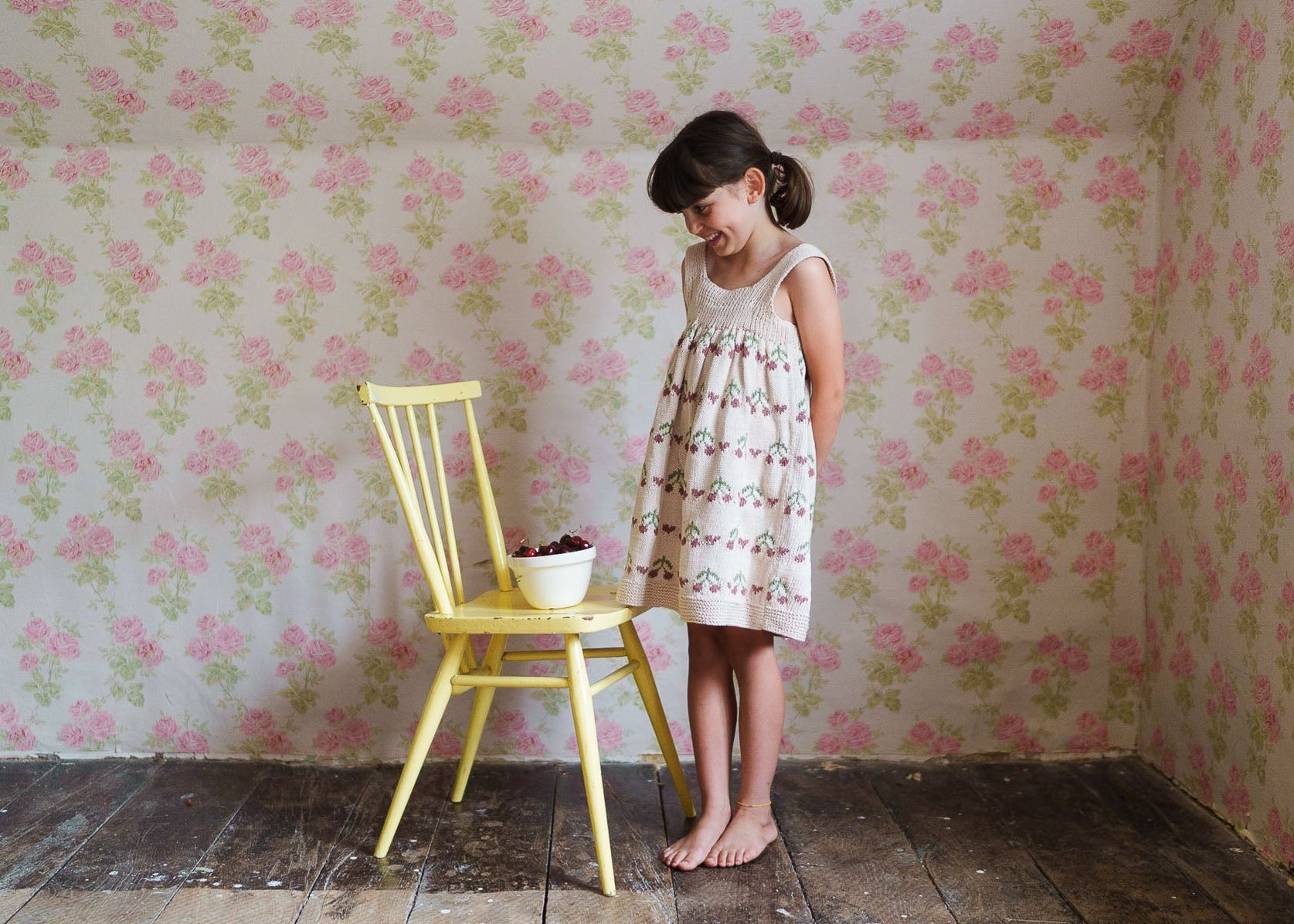 A young girl wearing a vintage-style, hand-knitted pinafore dress made from organic cotton, standing barefoot next to a yellow wooden chair with a bowl of cherries, in a room with floral wallpaper and rustic wooden floor.