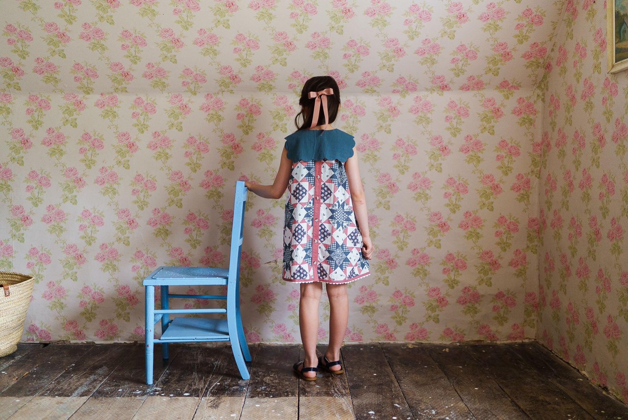Young girl wearing a vintage-style organic cotton dress with a scalloped blue collar, standing beside a blue wooden chair in a floral wallpapered room.