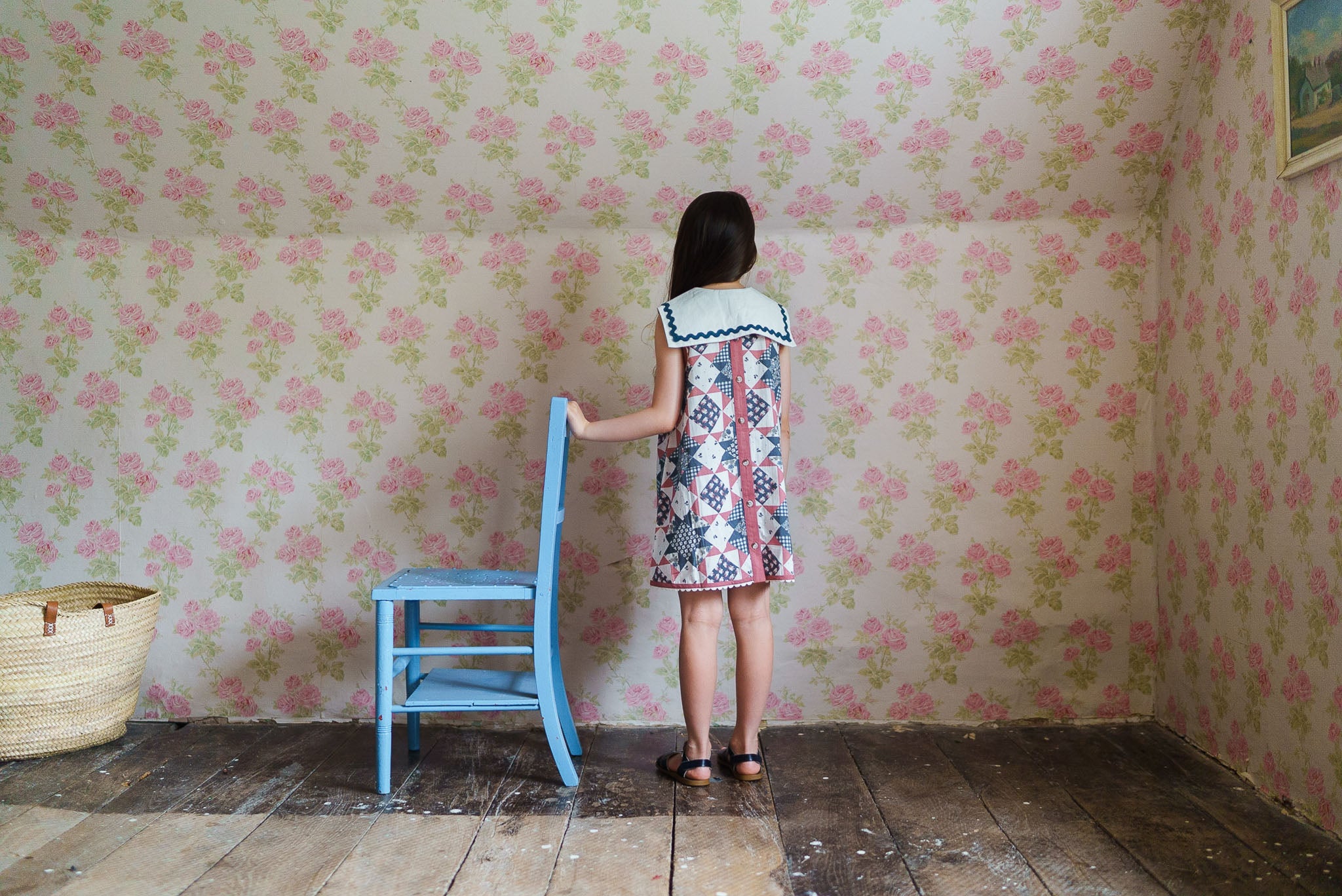 Girl wearing a sustainable vintage style dress with a sailor collar made from organic cotton, standing by a blue wooden chair in a rustic room with floral wallpaper.