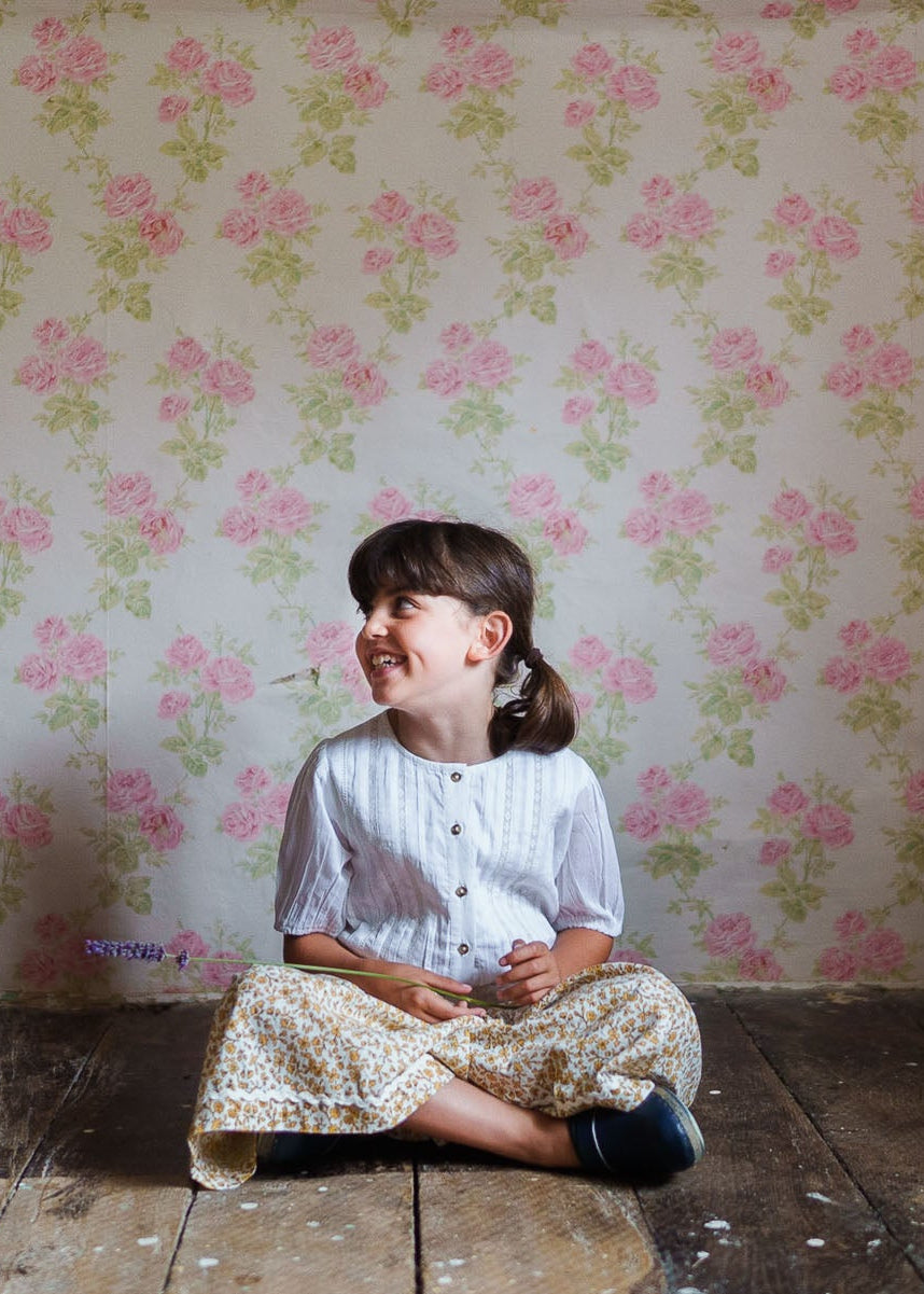 Smiling young girl sitting on wooden floor wearing an off-white organic cotton blouse with buttons, paired with floral vintage-style skirt against a rose-patterned wall.