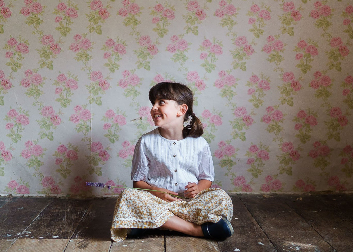 Smiling child sitting cross-legged on a rustic wooden floor wearing vintage-style floral culottes and a soft white blouse, showcasing sustainable organic children's clothing.